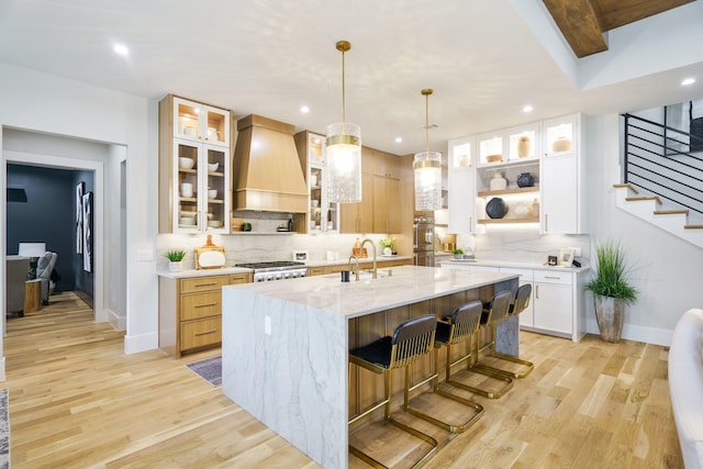 kitchen featuring premium range hood, a breakfast bar, light stone counters, a kitchen island with sink, and white cabinets