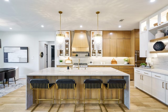 kitchen featuring decorative light fixtures, white cabinets, custom exhaust hood, a kitchen island with sink, and light stone counters