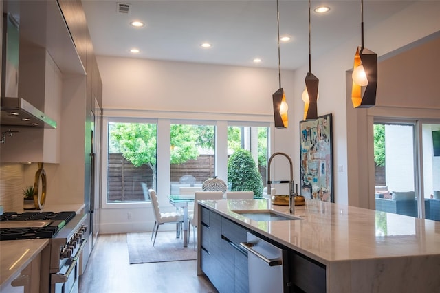 kitchen featuring light stone countertops, a kitchen island with sink, sink, pendant lighting, and wall chimney exhaust hood