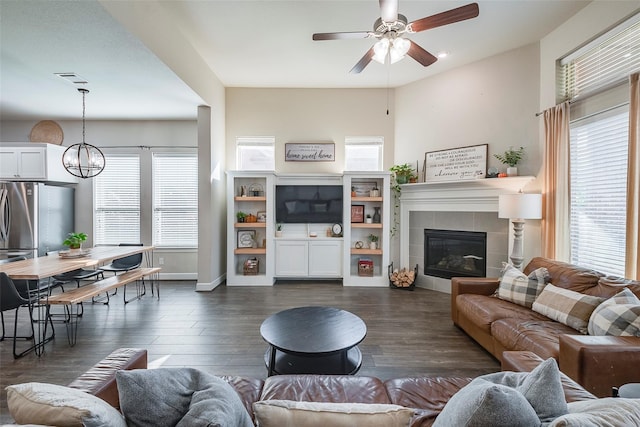 living room with ceiling fan with notable chandelier, dark wood-type flooring, a tile fireplace, and a healthy amount of sunlight