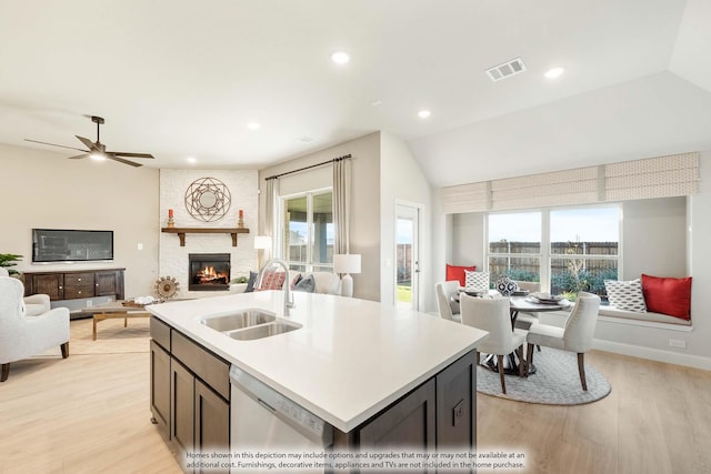 kitchen featuring sink, a large fireplace, a kitchen island with sink, stainless steel dishwasher, and light hardwood / wood-style flooring