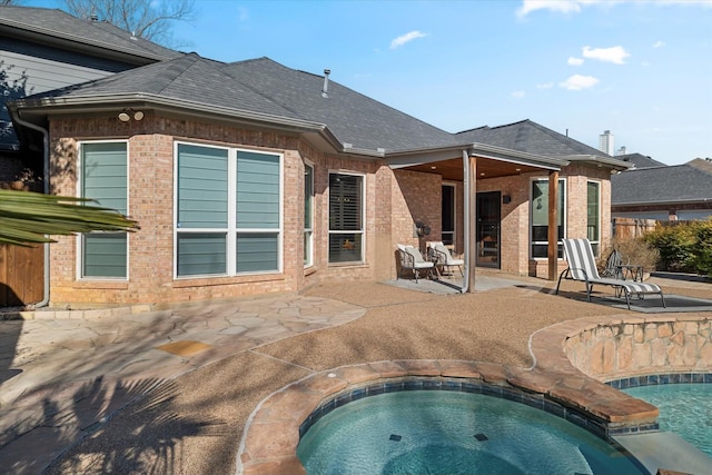 rear view of house with an in ground hot tub, brick siding, a patio area, and a shingled roof