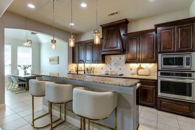 kitchen featuring custom exhaust hood, a kitchen bar, stainless steel appliances, light stone counters, and a center island with sink