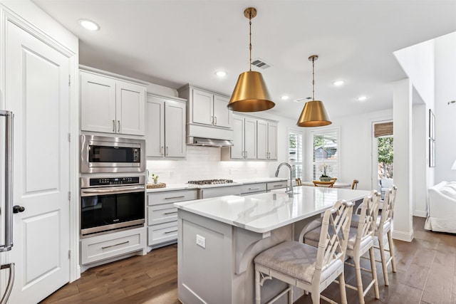 kitchen featuring appliances with stainless steel finishes, a kitchen island with sink, dark wood-type flooring, and tasteful backsplash