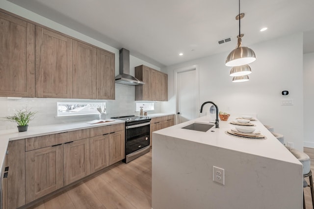 kitchen featuring a kitchen island with sink, sink, wall chimney range hood, pendant lighting, and stainless steel range with electric cooktop