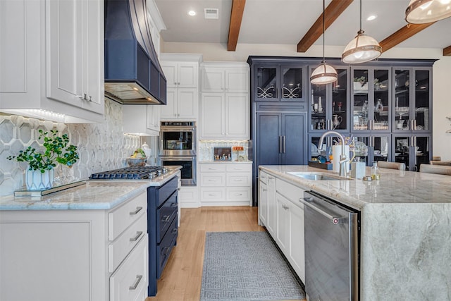 kitchen with custom exhaust hood, hanging light fixtures, sink, stainless steel appliances, and a center island with sink