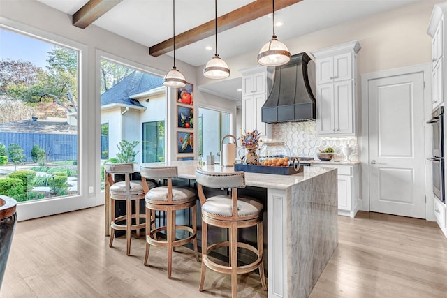 kitchen with pendant lighting, white cabinetry, premium range hood, a kitchen island with sink, and light stone counters