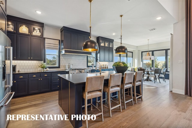 kitchen with high quality fridge, backsplash, hanging light fixtures, a kitchen island with sink, and light hardwood / wood-style flooring