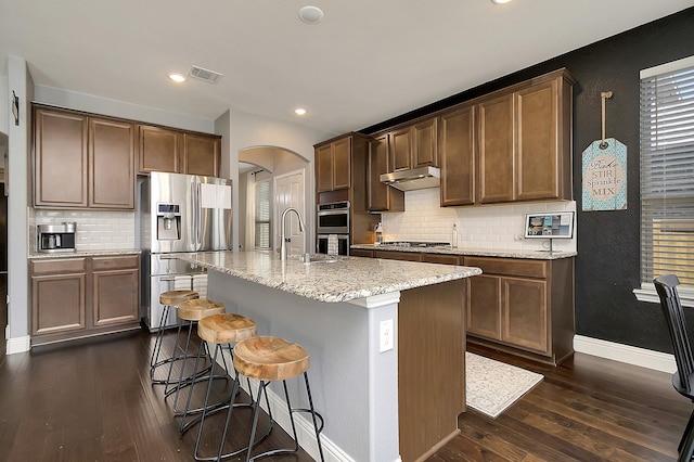 kitchen with sink, dark wood-type flooring, stainless steel appliances, light stone countertops, and a center island with sink