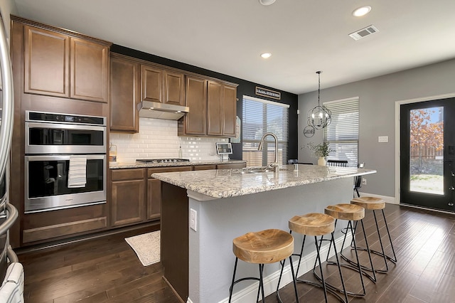 kitchen featuring a kitchen island with sink, stainless steel appliances, light stone counters, decorative backsplash, and decorative light fixtures