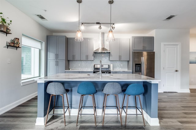 kitchen with pendant lighting, appliances with stainless steel finishes, gray cabinets, wall chimney range hood, and a center island with sink