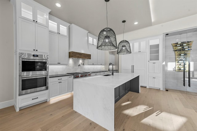 kitchen with pendant lighting, custom range hood, white cabinetry, a center island with sink, and stainless steel appliances