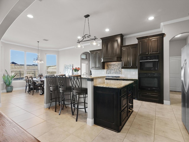 kitchen featuring premium range hood, tasteful backsplash, dark brown cabinets, an island with sink, and pendant lighting