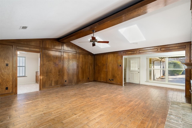 unfurnished living room featuring light wood-type flooring, ceiling fan, lofted ceiling with skylight, and wood walls