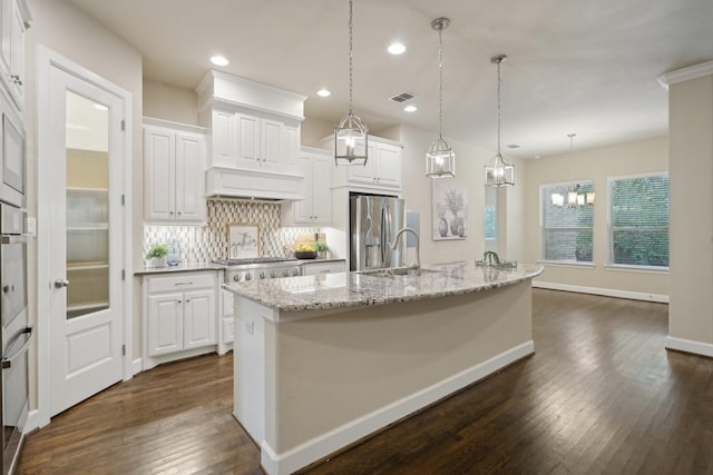 kitchen with white cabinets, appliances with stainless steel finishes, decorative light fixtures, decorative backsplash, and a kitchen island with sink