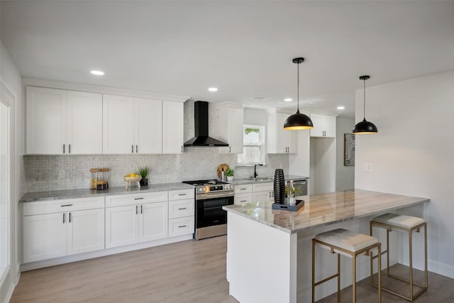 kitchen with hanging light fixtures, wall chimney range hood, white cabinets, stainless steel gas stove, and light stone counters