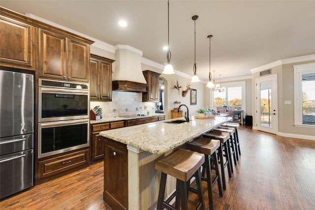 kitchen with stainless steel appliances, custom range hood, backsplash, a kitchen island with sink, and a sink