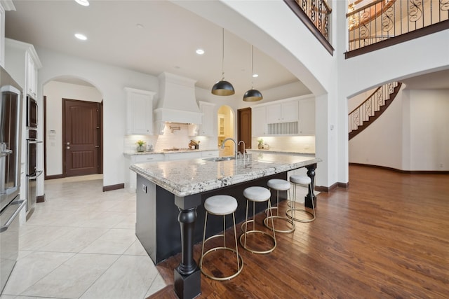 kitchen with premium range hood, pendant lighting, white cabinets, light stone counters, and a center island with sink