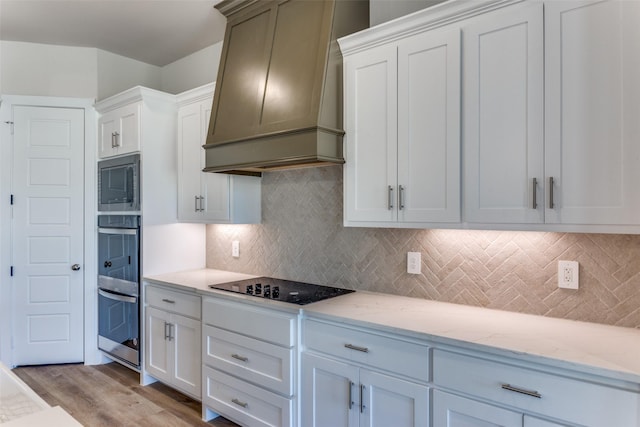 kitchen featuring light stone countertops, premium range hood, light wood finished floors, decorative backsplash, and appliances with stainless steel finishes