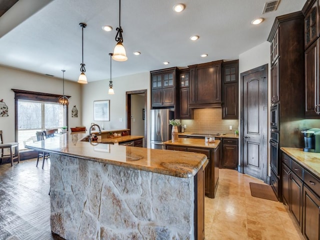 kitchen with appliances with stainless steel finishes, custom range hood, dark brown cabinets, decorative light fixtures, and an island with sink