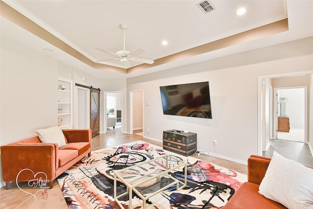 living room with built in shelves, ceiling fan, crown molding, a tray ceiling, and a barn door