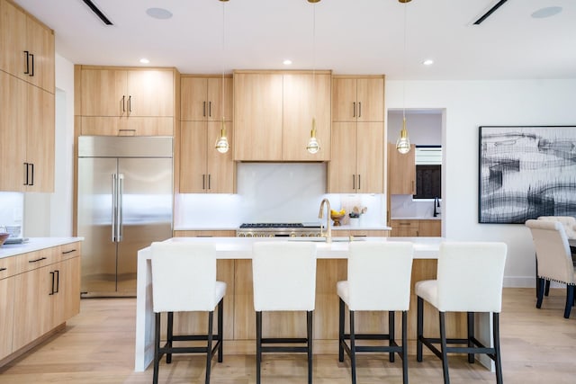 kitchen with decorative light fixtures, a kitchen island with sink, built in fridge, and light brown cabinetry