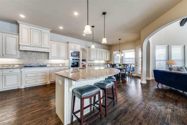 kitchen with light stone countertops, hanging light fixtures, stainless steel appliances, and a kitchen island with sink