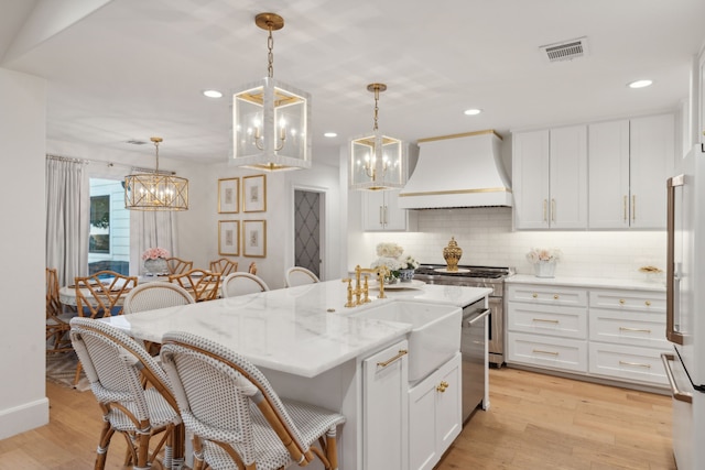 kitchen featuring decorative light fixtures, premium range hood, white cabinets, and a center island with sink