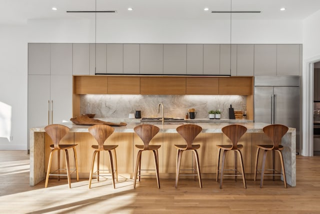 kitchen featuring built in refrigerator, light stone counters, a kitchen bar, decorative backsplash, and light wood-type flooring