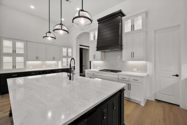 kitchen with premium range hood, decorative light fixtures, stainless steel gas stovetop, white cabinetry, and a center island with sink