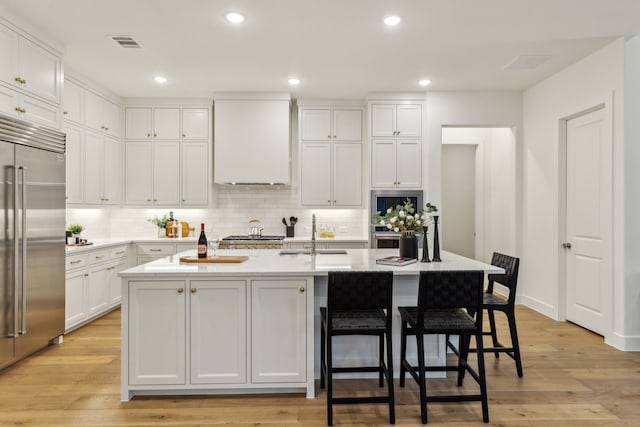 kitchen with sink, a center island with sink, appliances with stainless steel finishes, white cabinets, and wall chimney range hood