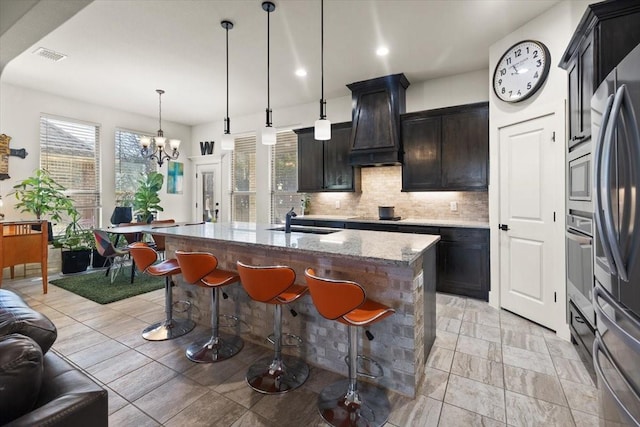 kitchen with decorative light fixtures, a kitchen island with sink, light stone counters, stainless steel appliances, and custom range hood