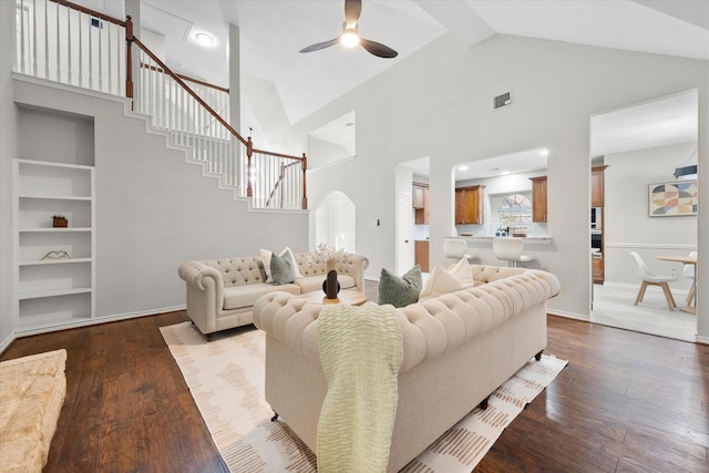living room featuring high vaulted ceiling, built in shelves, a ceiling fan, dark wood-style floors, and stairway