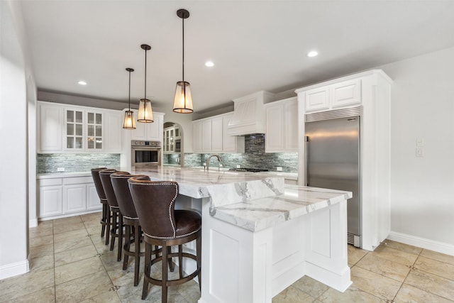 kitchen featuring custom exhaust hood, white cabinetry, a large island with sink, pendant lighting, and stainless steel appliances