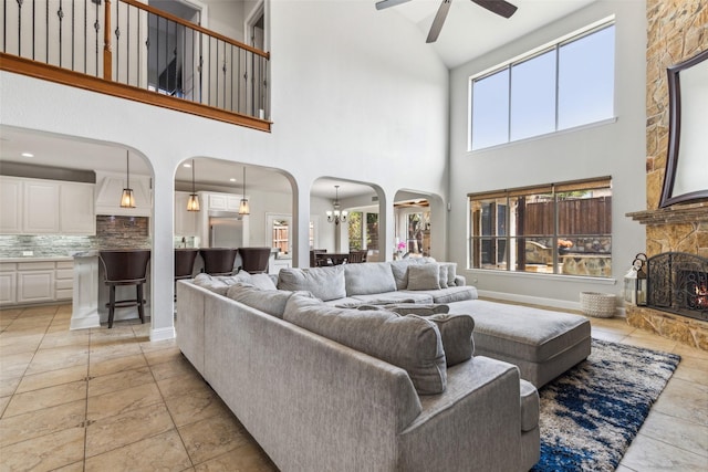 living room with a stone fireplace, ceiling fan with notable chandelier, a wealth of natural light, and a high ceiling