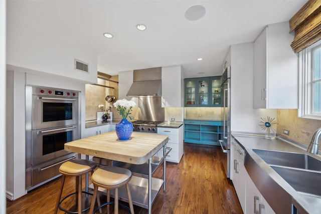 kitchen featuring wall chimney range hood, decorative backsplash, stainless steel appliances, and white cabinets