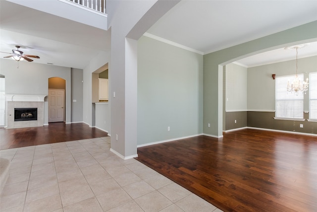 unfurnished living room featuring a tiled fireplace, light tile patterned flooring, ceiling fan with notable chandelier, and crown molding