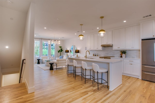 kitchen with white cabinetry, decorative light fixtures, light hardwood / wood-style flooring, stainless steel refrigerator, and a kitchen island with sink