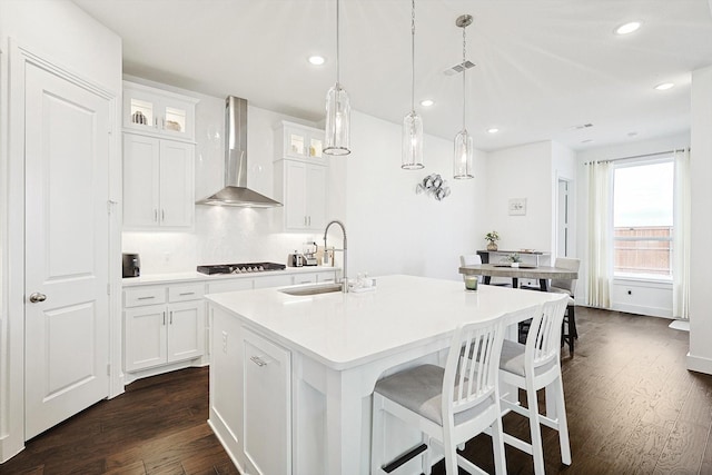 kitchen featuring sink, white cabinetry, decorative light fixtures, a center island with sink, and wall chimney range hood