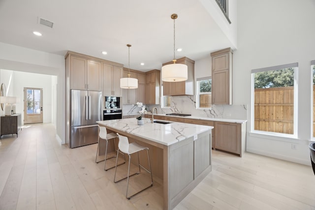 kitchen featuring appliances with stainless steel finishes, hanging light fixtures, light stone counters, a center island with sink, and decorative backsplash