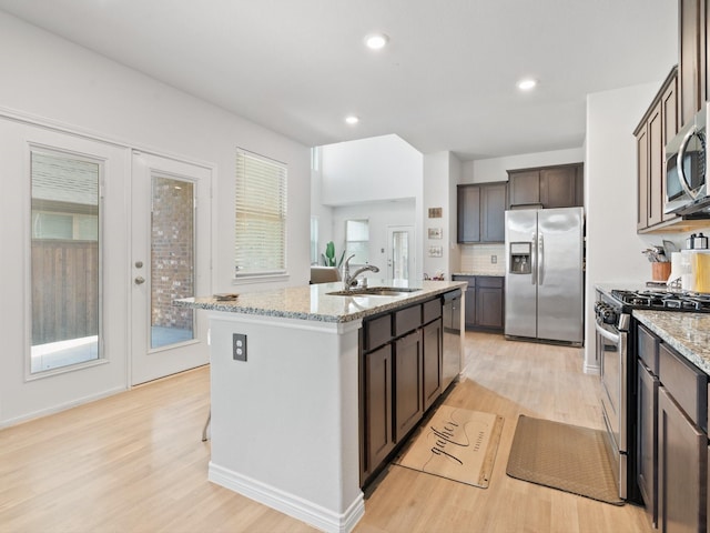 kitchen featuring sink, stainless steel appliances, dark brown cabinetry, light stone countertops, and a center island with sink