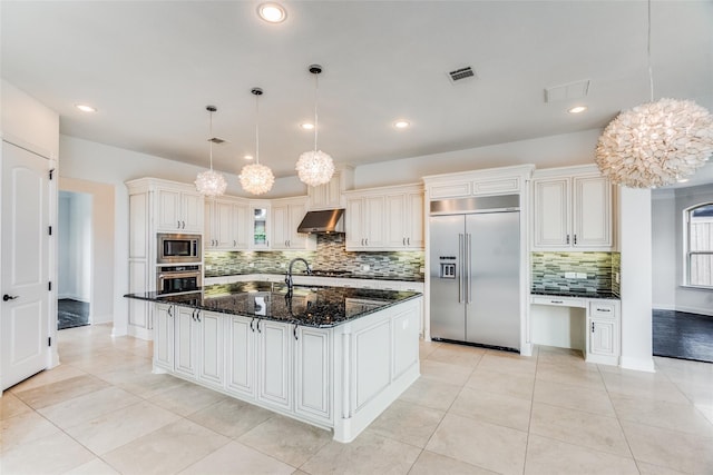 kitchen with built in appliances, decorative light fixtures, light tile patterned floors, an island with sink, and dark stone counters