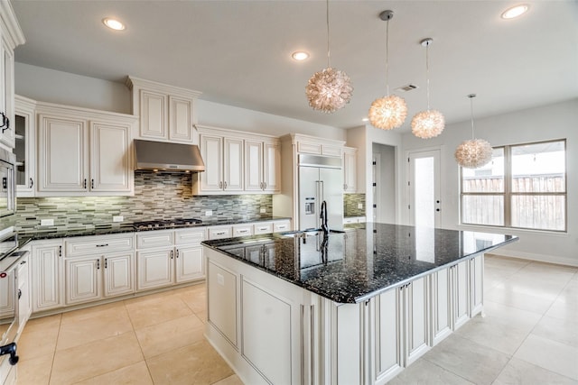 kitchen with stainless steel appliances, extractor fan, a kitchen island with sink, and dark stone countertops