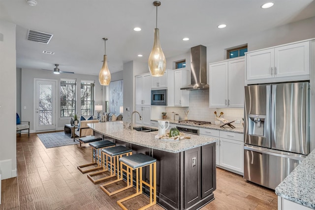 kitchen with appliances with stainless steel finishes, pendant lighting, an island with sink, white cabinets, and wall chimney range hood