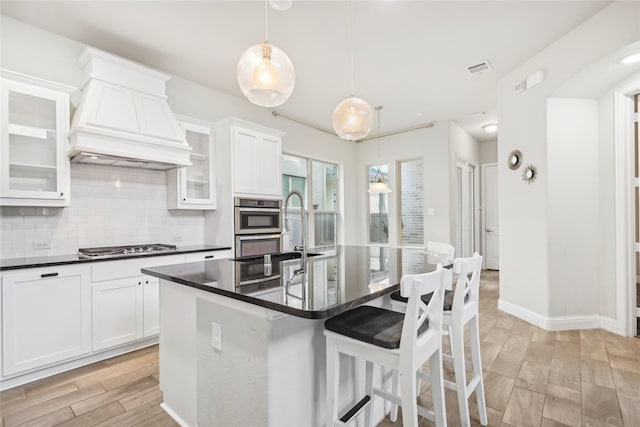 kitchen with hanging light fixtures, an island with sink, custom range hood, white cabinets, and backsplash