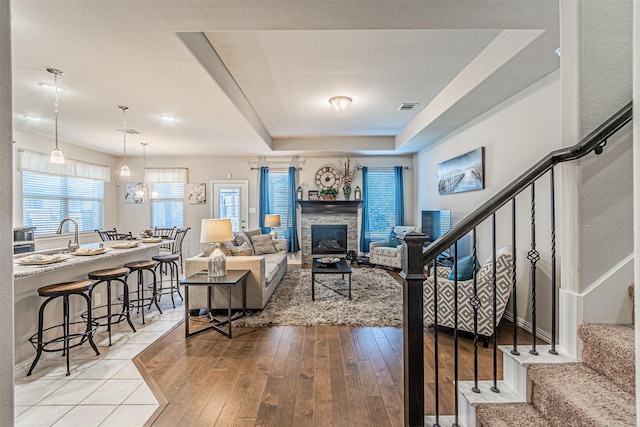 living room with an inviting chandelier, a stone fireplace, a raised ceiling, and light wood-type flooring