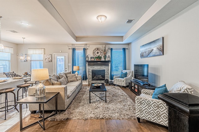 living room with a raised ceiling, a stone fireplace, hardwood / wood-style floors, and a notable chandelier