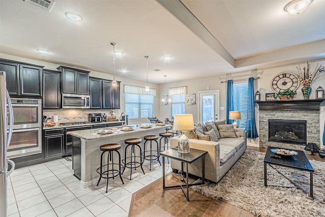 kitchen featuring appliances with stainless steel finishes, pendant lighting, a kitchen breakfast bar, a kitchen island with sink, and light stone countertops