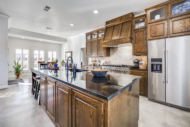 kitchen featuring premium range hood, sink, high end refrigerator, dark stone countertops, and a kitchen island with sink