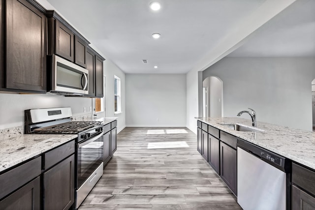 kitchen with dark brown cabinetry, sink, light wood-type flooring, stainless steel appliances, and light stone countertops